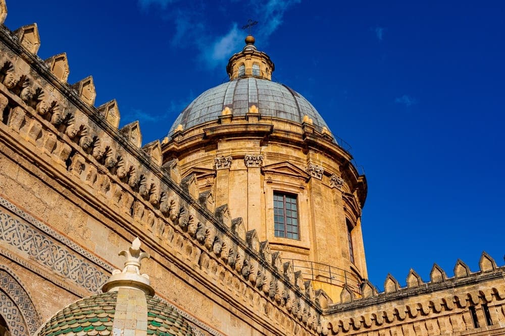 The Palermo Cathedral in Palermo, Italy, featuring ornate domes and detailed stonework against a bright blue sky.