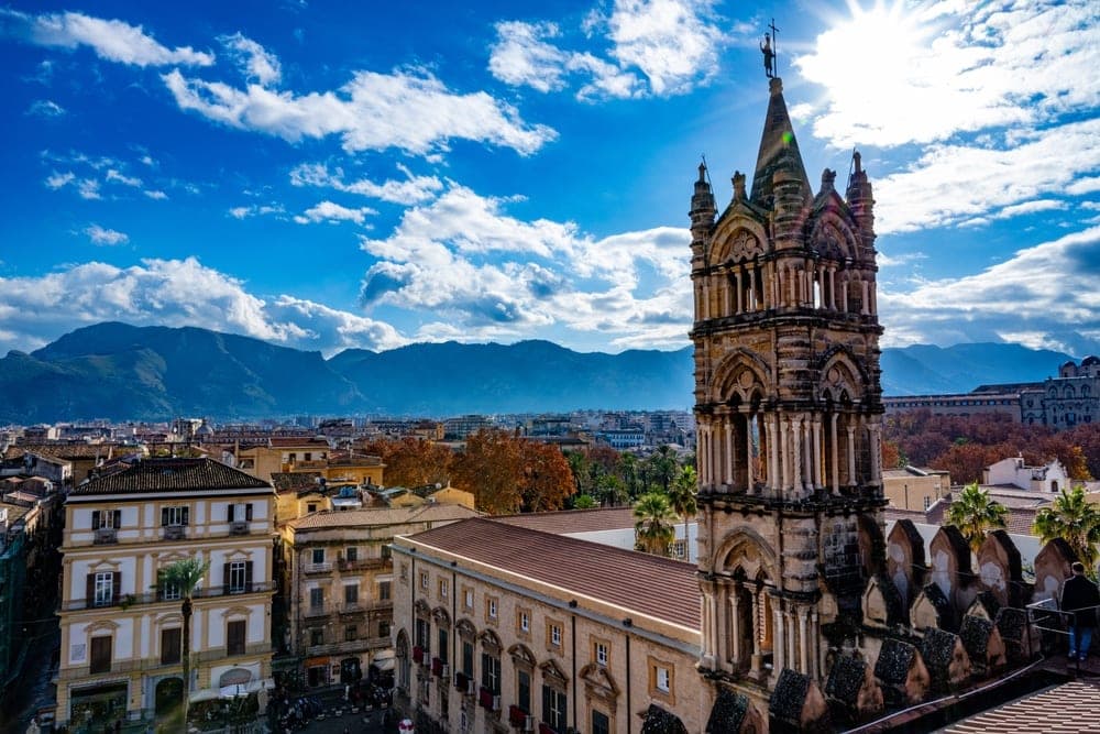 The detailed stone bell tower of Palermo Cathedral in Italy stands tall against a bright blue sky with clouds, overlooking the cityscape of Palermo and distant mountains.