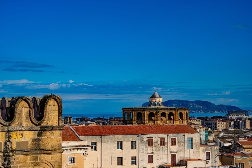Elevated view of Palermo, Italy, showing historic stone architecture, a domed building, and the Mediterranean Sea under a bright blue sky.