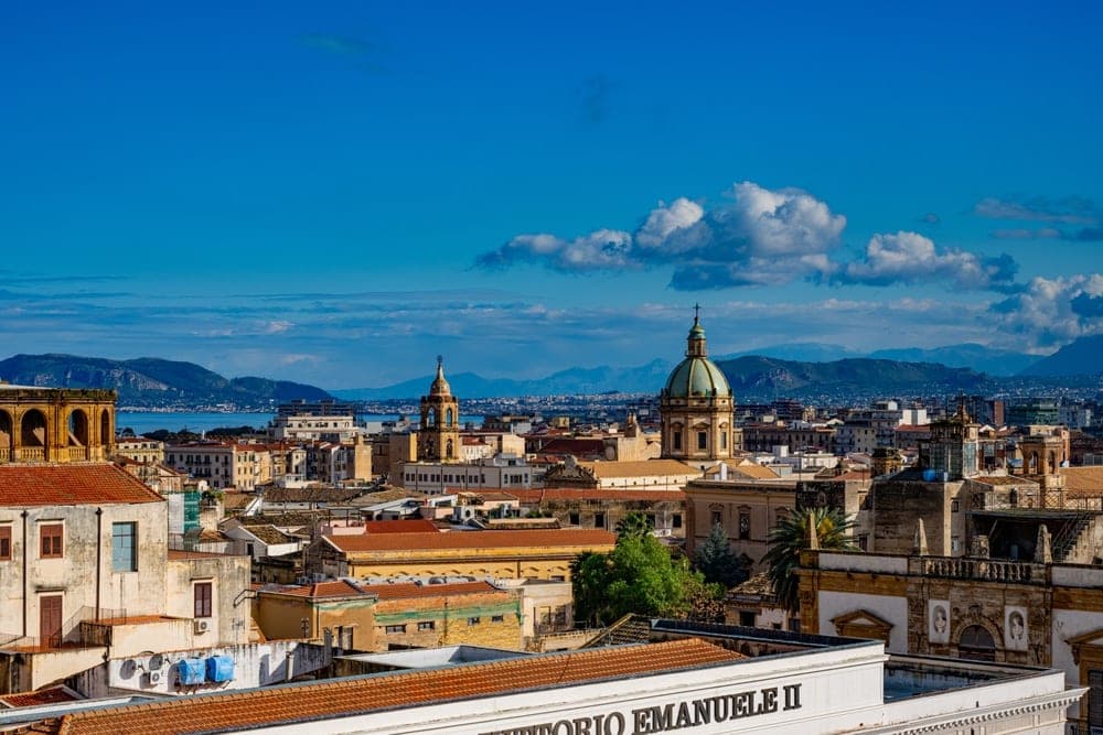Panoramic view of Palermo, Italy, with historic buildings, domes, and distant mountains under a bright blue sky.