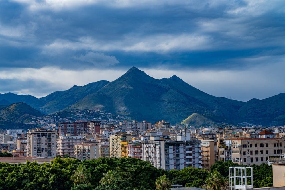 View of Palermo, Italy, with dense urban buildings in the foreground and dramatic, dark mountains under a cloudy sky.