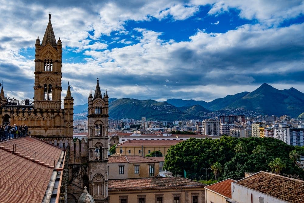 Panoramic view of Palermo, Italy, featuring the spires of the Palermo Cathedral in the foreground and mountains in the background.