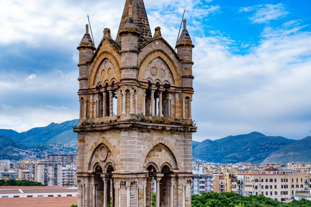 The medieval bell tower of Palermo Cathedral stands tall against a cloudy blue sky, overlooking the cityscape of Palermo, Italy.