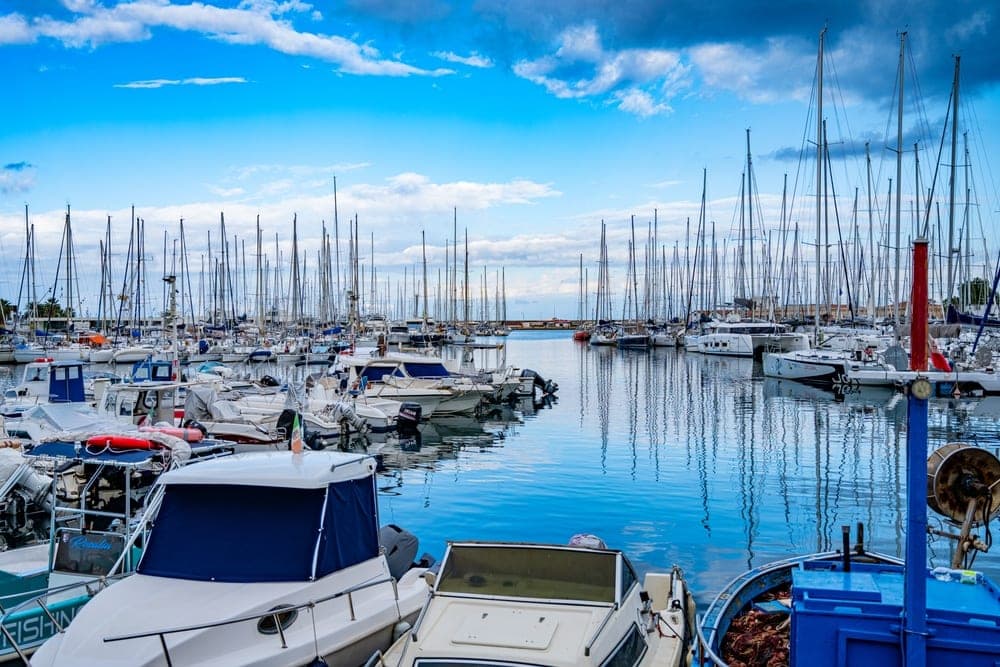 A busy marina in Palermo, Italy, filled with sailboats and motorboats under a bright blue sky with white clouds.