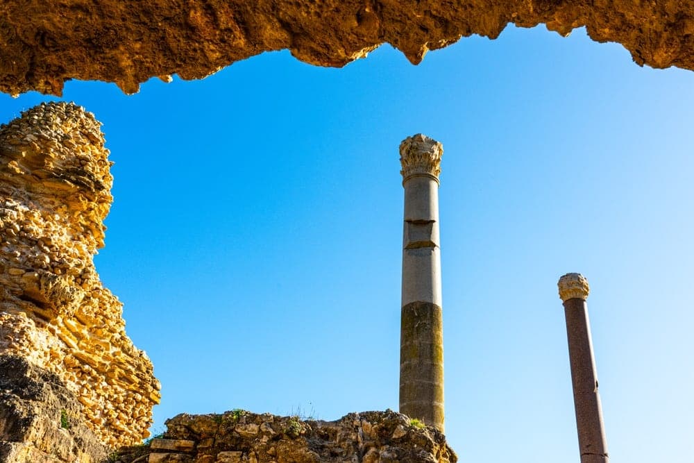 Ancient Roman ruins in Tunis, Tunisia, with two prominent columns against a clear blue sky, framed by a rocky overhang.