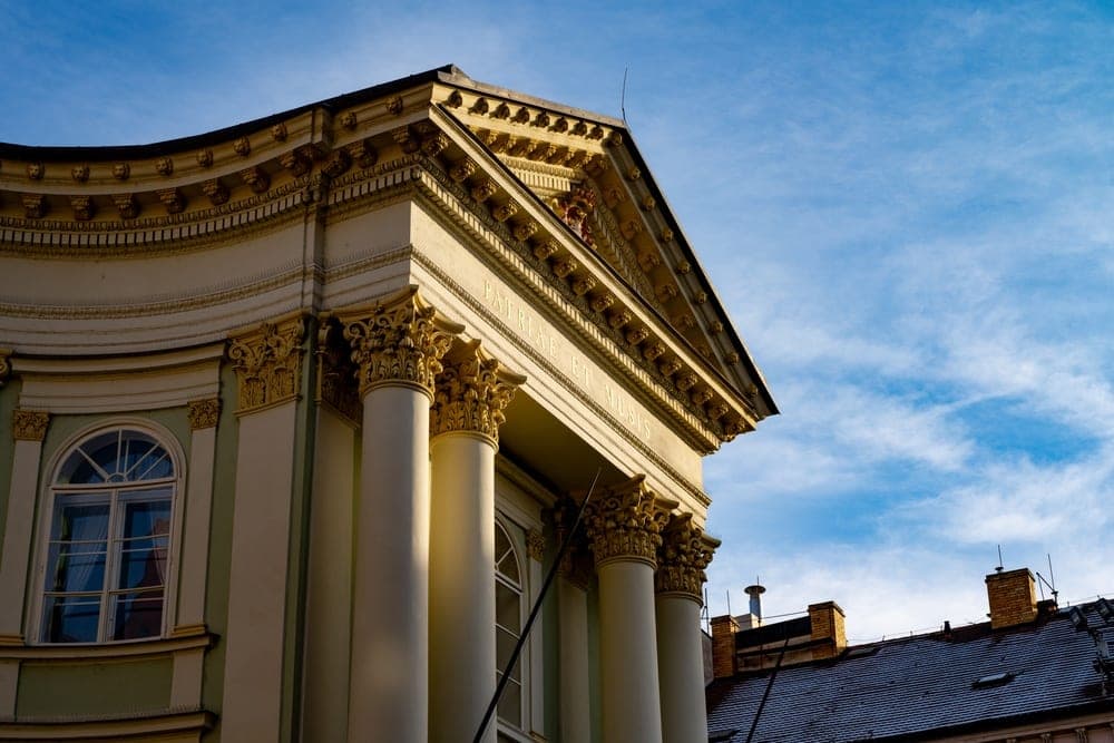 The grand facade of a historical building with white Corinthian columns and gold leaf details in Prague, Czech Republic, under a bright blue sky.