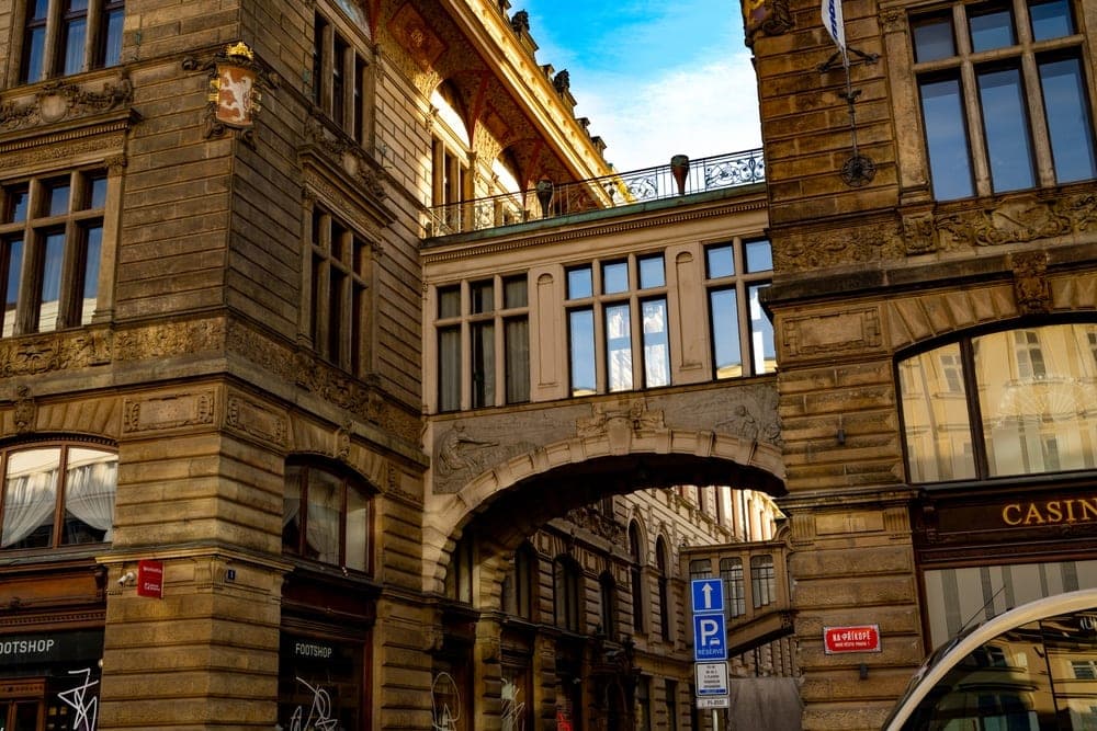 Ornate historical buildings in Prague, Czech Republic, connected by an arched bridge, under a bright blue sky.