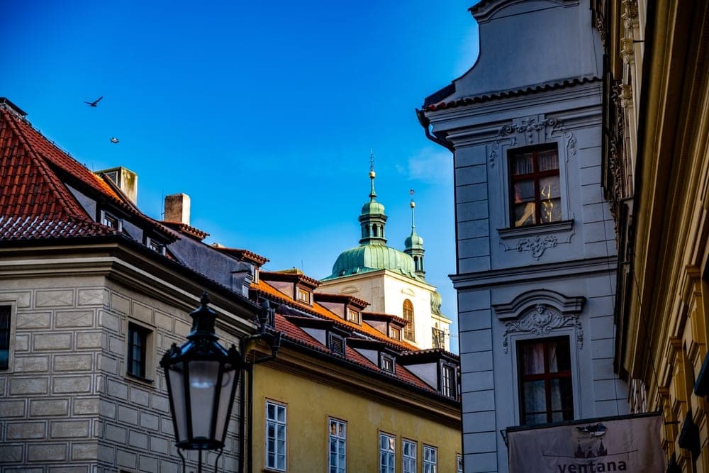 Historic buildings and a green-domed church tower in Prague, Czech Republic, under a bright blue sky.