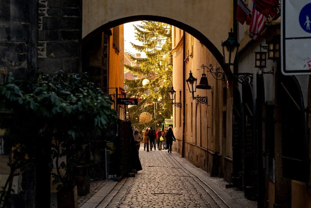 A view through an archway in Prague, Czechia, revealing a festive Christmas tree and cobblestone street.