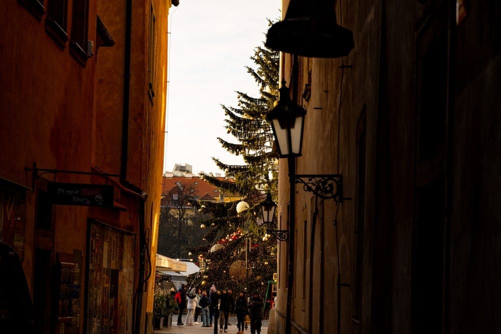 A festive Christmas tree stands at the end of a narrow, cobblestone alley in Prague, Czech Republic, illuminated by evening lights.
