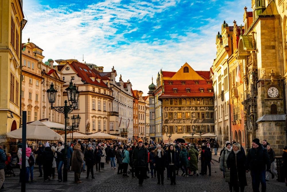 A bustling Staroměstské Náměstí (Old Town Square) in Prague, Czech Republic, with crowds of people, ornate historic buildings, and a bright blue sky with clouds. Golden light illuminates the scene.