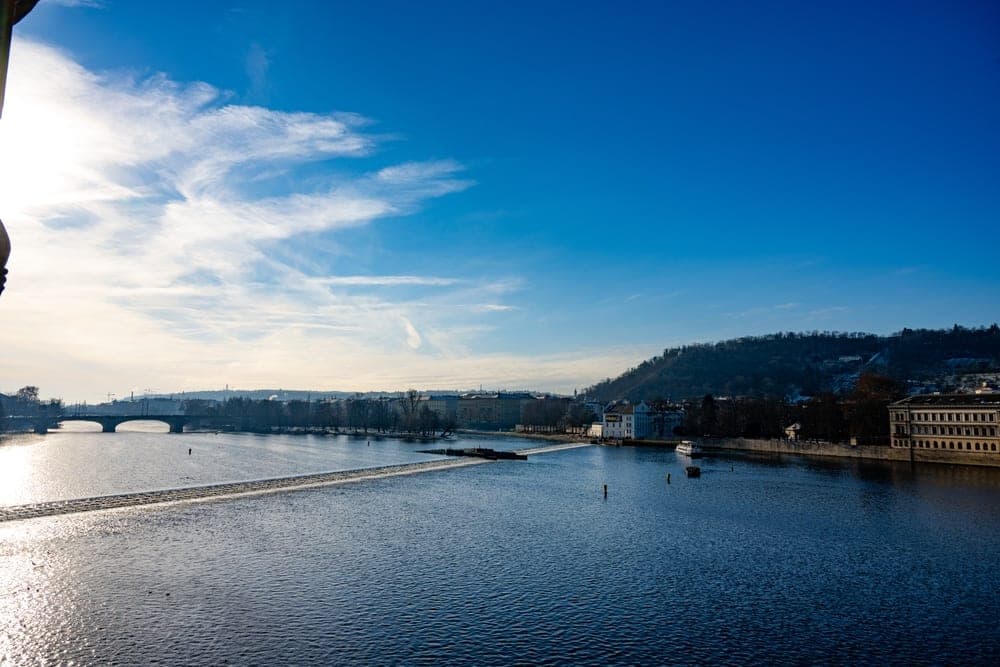 The Vltava River in Prague, Czech Republic, with a weir and a bridge under a bright blue sky with wispy clouds.