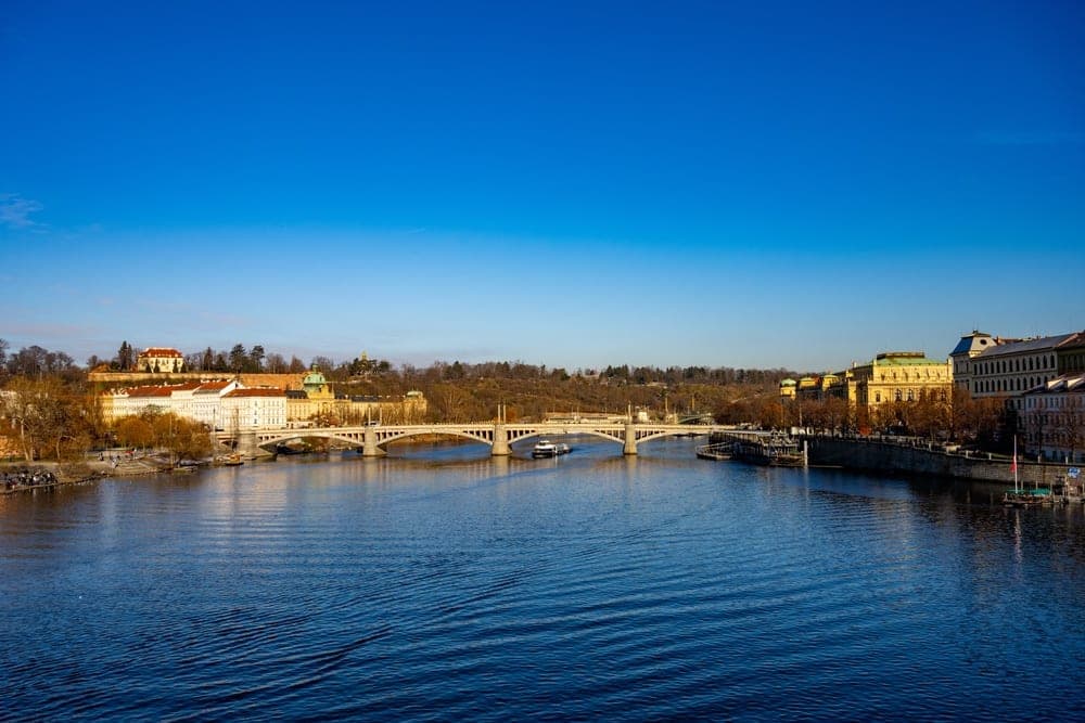The Mánes Bridge spans the Vltava River in Prague, Czech Republic, with historic buildings on both banks under a clear blue sky.