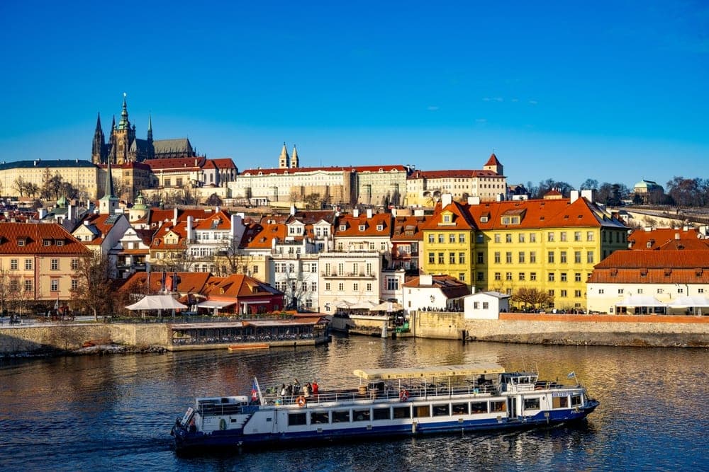 View of Prague, Czech Republic, with Prague Castle and St. Vitus Cathedral overlooking the Vltava River and Old Town architecture, with a passenger boat on the water.