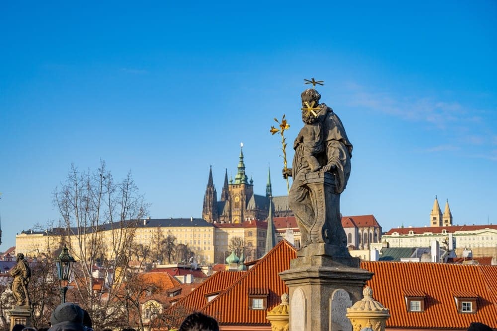 A statue on Charles Bridge in Prague, Czech Republic, with Prague Castle in the background under a clear blue sky.