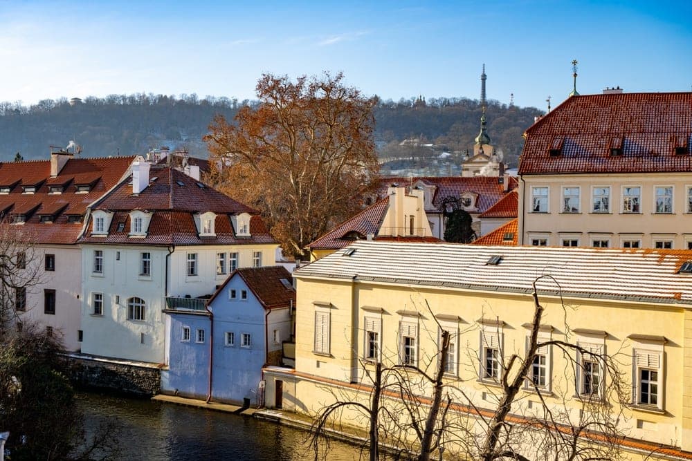 A winter view of Prague, Czech Republic, with snow-dusted terracotta roofs and historic buildings along a canal, under a clear blue sky.