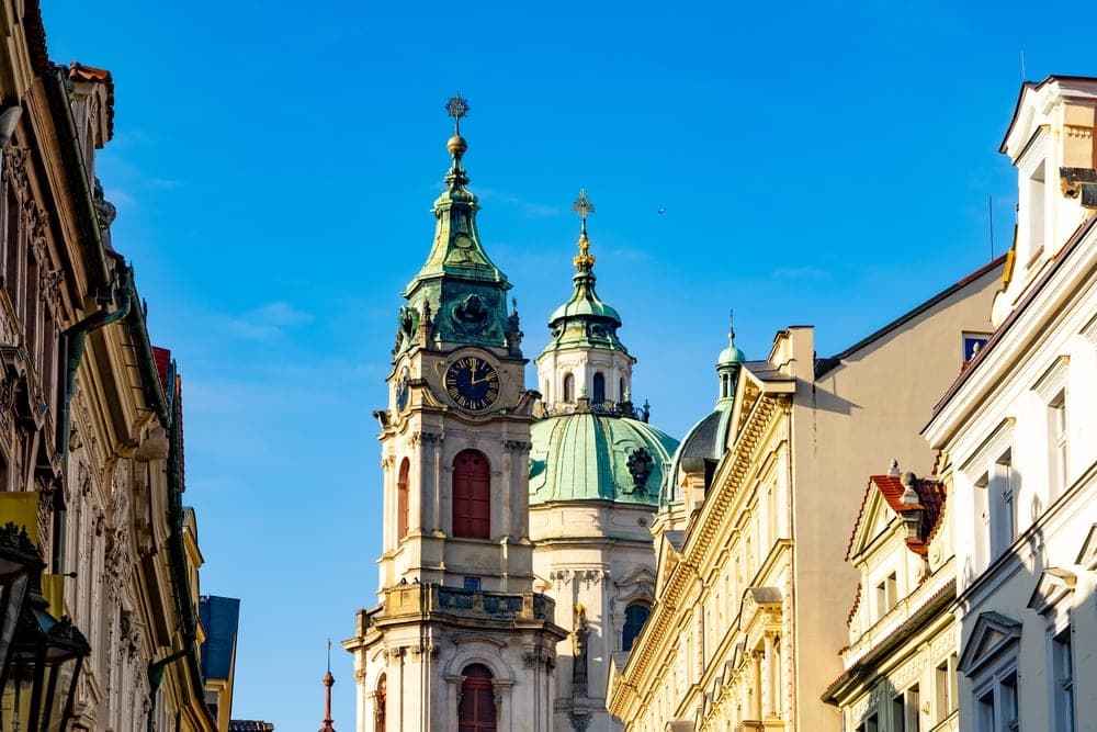 A view up a street in Prague, Czechia, towards St. Nicholas Church with its prominent clock tower and green domes, under a bright blue sky.