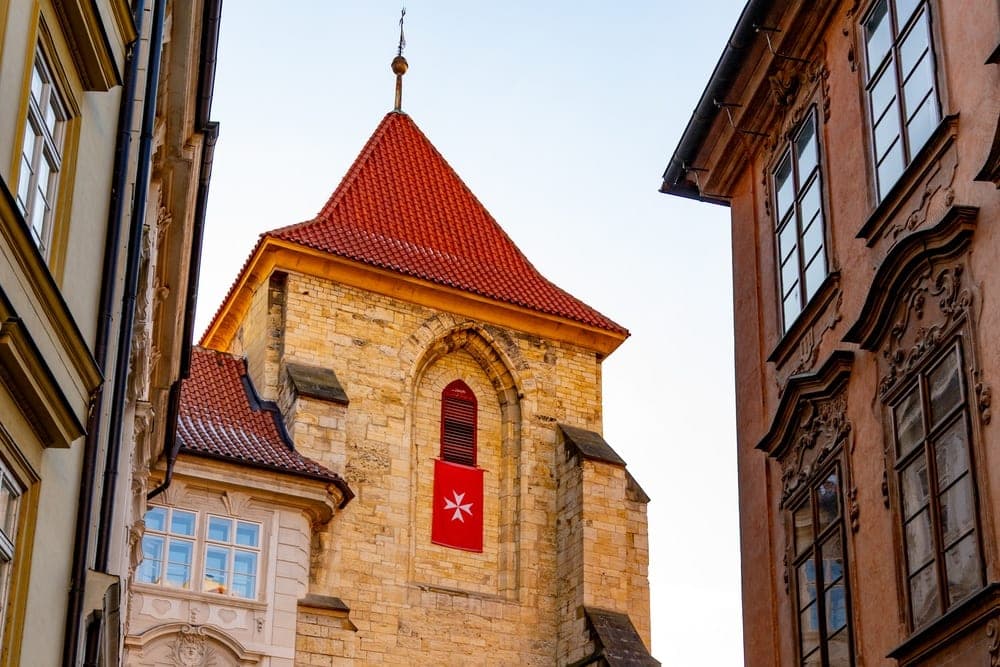 A cobblestone street in Prague, Czech Republic, leading towards a stone church with a red roof and a prominent white Maltese cross banner.
