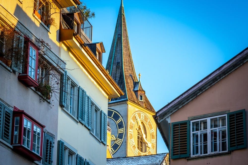 St. Peter's Church clock tower in Zürich, Switzerland, viewed between colourful buildings with green shutters.