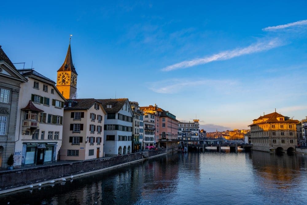 Historic buildings along the Limmat River in Zürich, Switzerland, bathed in the golden light of sunset, with a prominent clock tower.