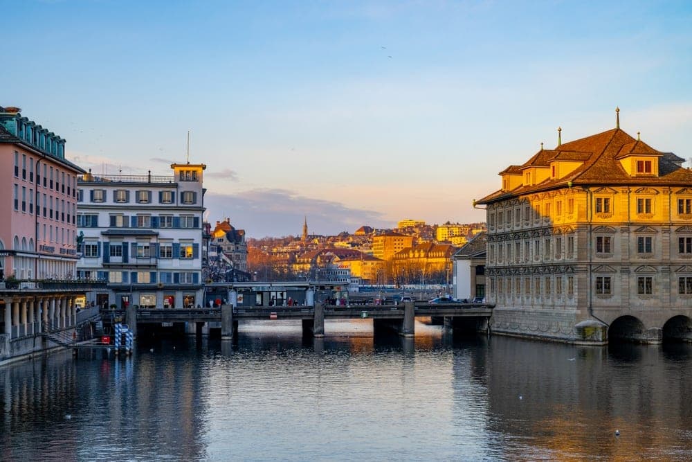 Golden hour sunset over the Limmat River in Zürich, Switzerland, with ornate buildings and a pedestrian bridge.