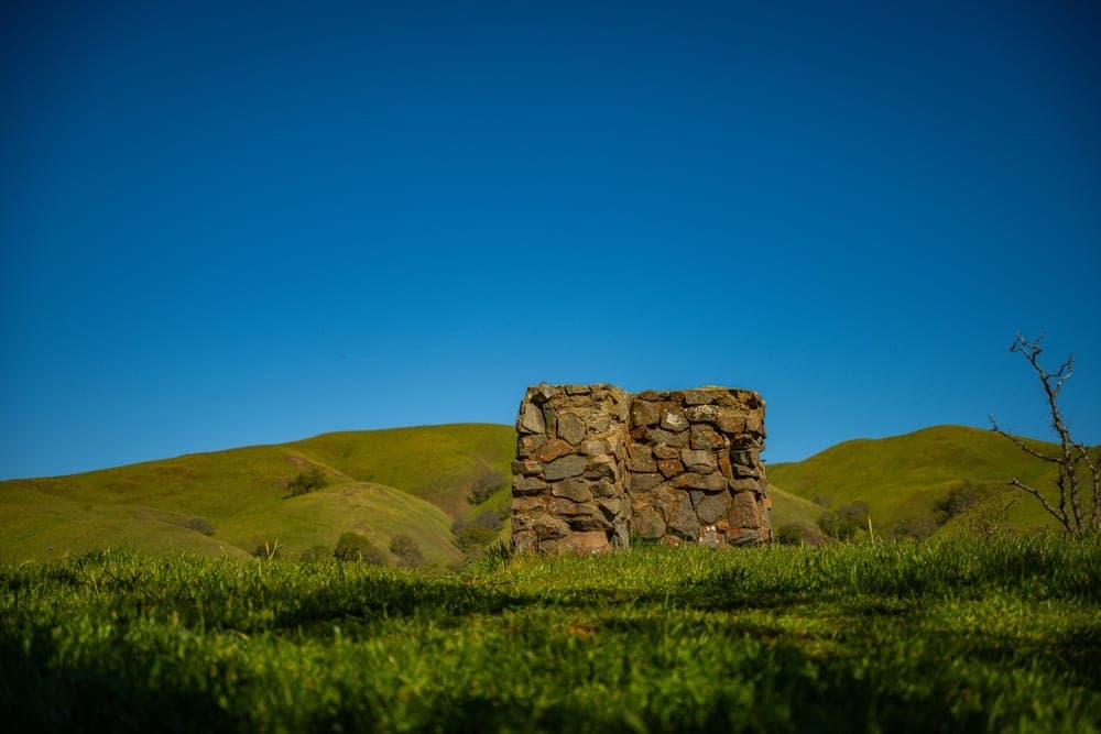 A weathered stone structure stands in a field of green grass, with rolling hills and a bright blue sky in Sunol, California, United States.