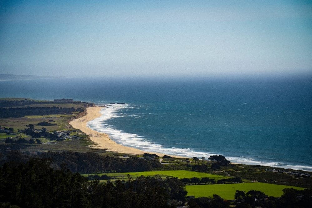 A sweeping view of the California coastline in San Francisco, United States, showing a sandy beach, blue ocean with waves, and green fields.