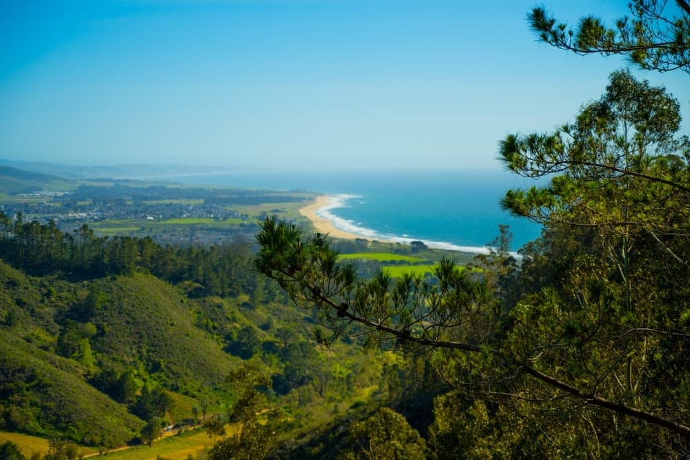 View of the coastline near El Granada, California, United States, with green hills in the foreground, a beach, and the Pacific Ocean under a clear blue sky.