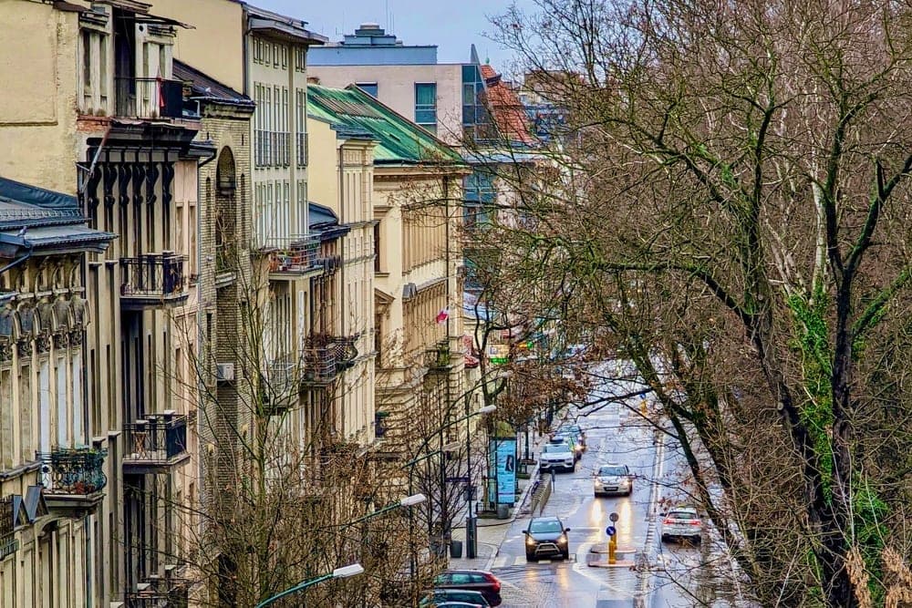 A wet street in Krakow, Poland, lined with old buildings and bare winter trees, with cars driving on the road.